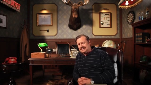 Image of "Dr.Mahogany" sitting in a leather chair with a moose head hanging on the wall behind him and other various decorations.